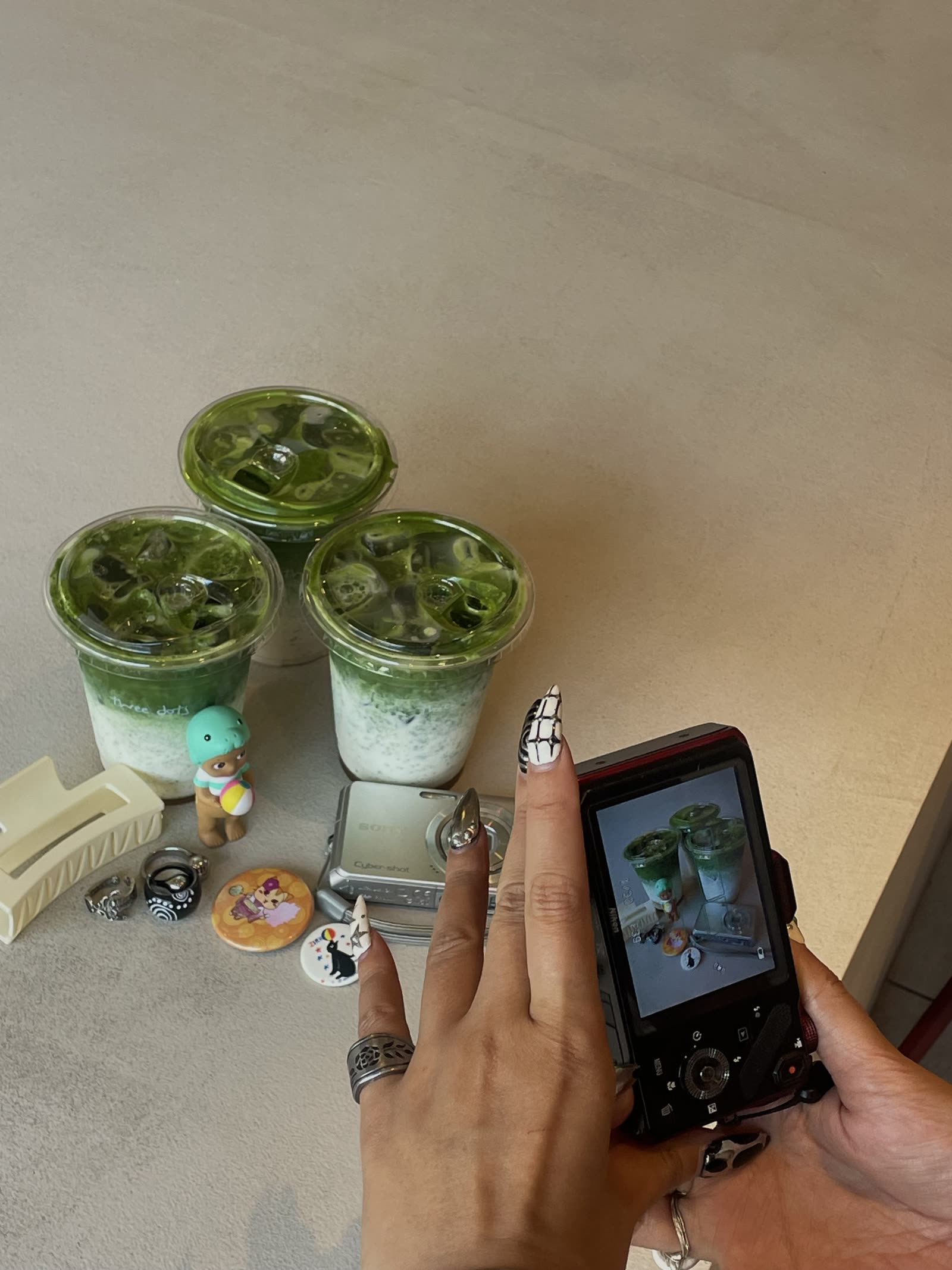 A hand with black-and-white checked and chrome nails holding a camera beside three iced matcha drinks