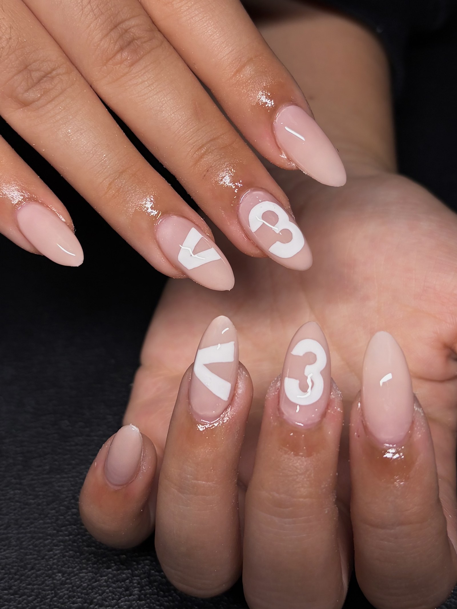 Milky nude almond nails with two accent nails carrying bold hand-painted white 'V' and '3' letter art